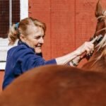 An older woman grooming a horse in front of a red stable