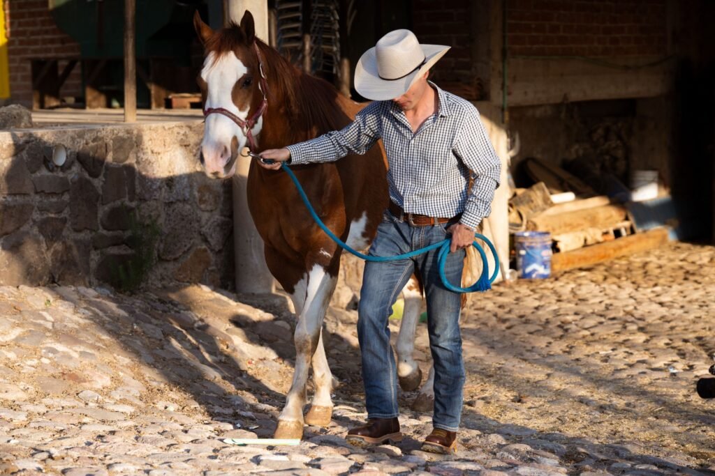 A cowboy leading a chestnut horse past a stable