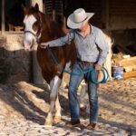 A cowboy leading a chestnut horse past a stable